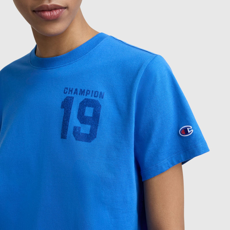 A person wears the Champion Grid Iron '19 Graphic T-Shirt featuring a bold "19" and "Champion" on the chest, C logo patch on the sleeve, sports-inspired style, set against a plain light gray background.