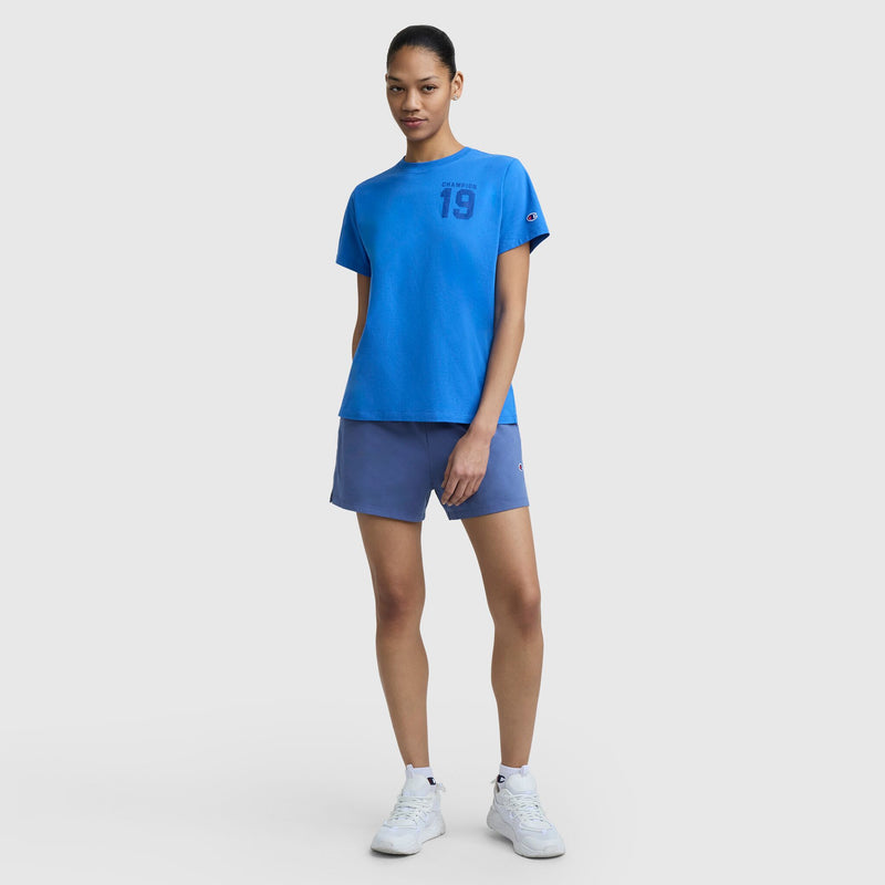 A woman stands against a light gray background in a vintage athletic look, wearing the Champion Grid Iron '19 Graphic T-Shirt with C Logo, blue athletic shorts, and white sneakers. Her hair is tied back as she looks confidently at the camera.