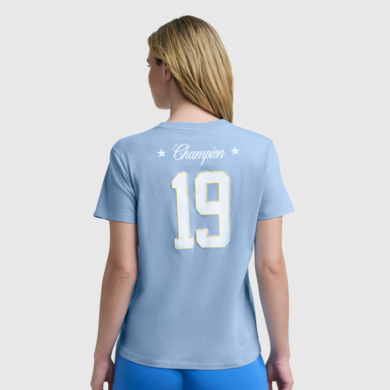 A person with long blonde hair is seen from behind, wearing a light blue Champion Gridiron Summer '19 Graphic T-Shirt with a white and yellow “19” and C Logo. The plain light background gives it a vintage sporty feel.