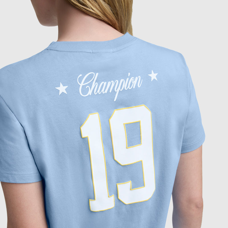 A person models the light blue Gridiron Summer '19 Graphic T-Shirt by Champion, featuring two stars and a C logo above a bold white and yellow 19 on the back, in vintage sport style, as they face away showing part of their face and upper body.