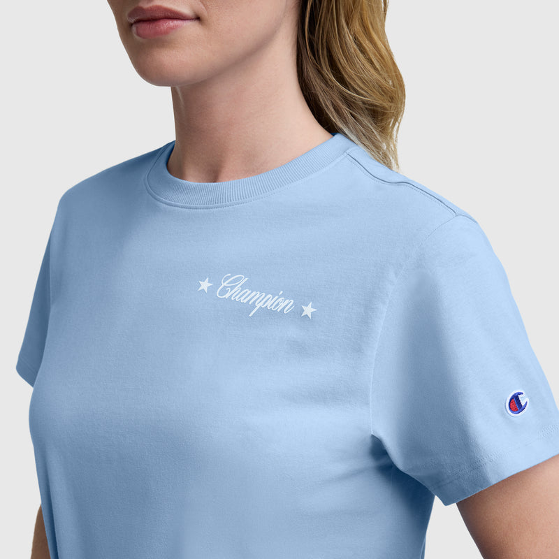 A woman wearing the Champion Gridiron Summer '19 Graphic T-Shirt, C Logo—a light blue tee featuring a white Champion graphic with two embroidered stars on the chest and a small logo patch on the left sleeve for vintage sport style.