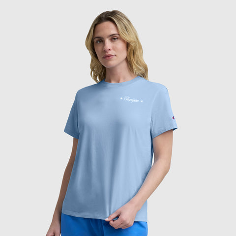 A woman with blonde hair wears a Champion Gridiron Summer '19 Graphic T-Shirt with the C logo, featuring two small white stars on the chest, evoking vintage sport style as she stands against a plain light gray background.