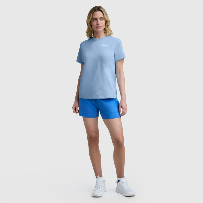 A woman stands facing forward, wearing a light blue Champion Gridiron Summer '19 Graphic T-Shirt with C Logo, bright blue shorts, and white sneakers. She has blonde hair and poses against a plain light background.