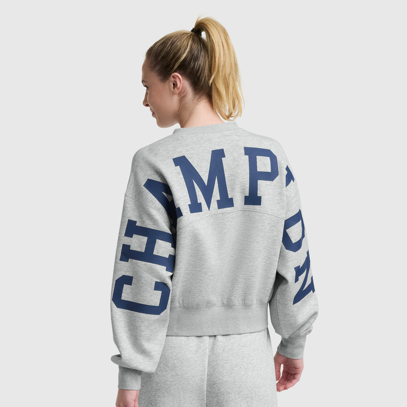 A woman with blonde hair in a ponytail is wearing a light gray Champion Endzone Loose Fit Crewneck with C Logo and sweatpants. The back and sleeve display the Champion HD graphic in bold blue. She is facing away from the camera.