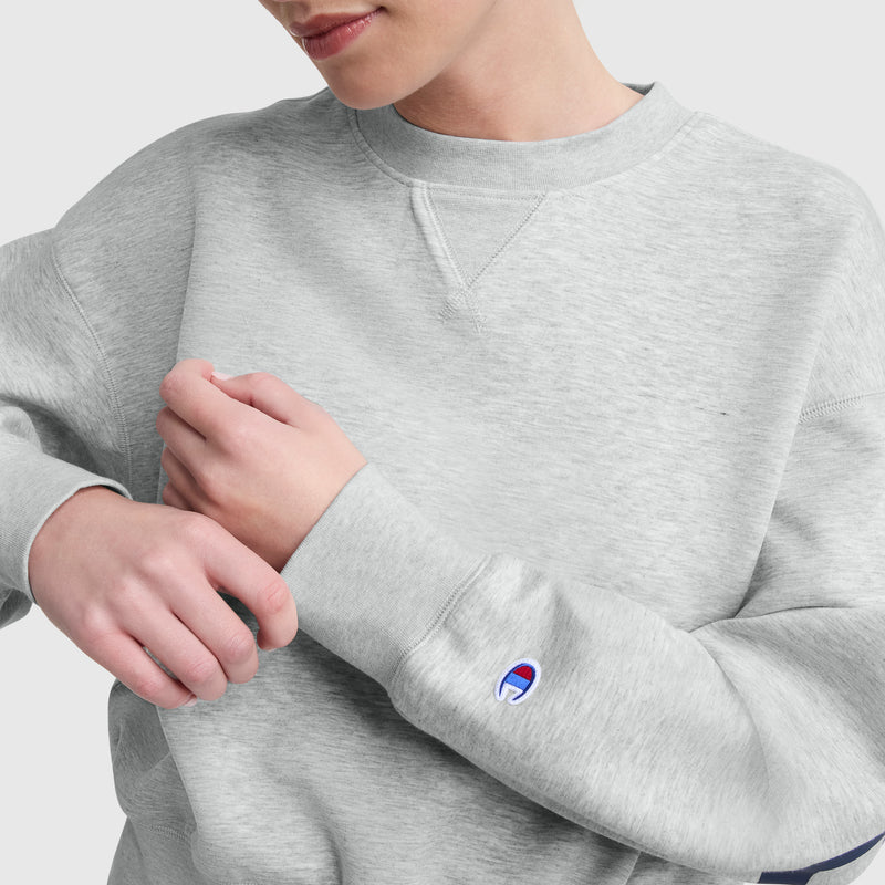 A person adjusts their cuff while wearing the Champion Endzone Loose Fit Crewneck, C Logo, in light gray scuba fabric, featuring a small blue and red Champion logo on the sleeve, set against a plain light background.