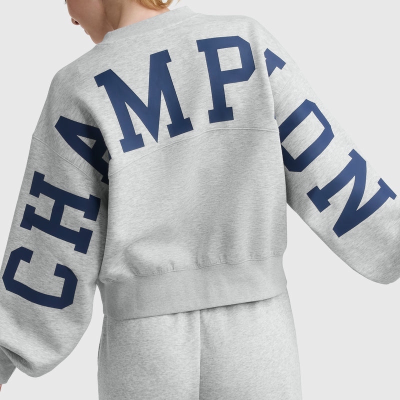 Person wearing a light gray Champion Endzone Loose Fit Crewneck with C Logo, shown from the back. Made from soft scuba fabric, this sweatshirt features a bold navy Champion HD graphic across the back and sleeves.