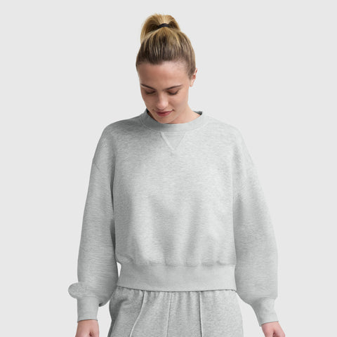 A woman with blonde hair in a ponytail wears a light gray Champion Endzone Loose Fit Crewneck, C Logo and matching sweatpants, looking down with a slight smile against a plain light background.