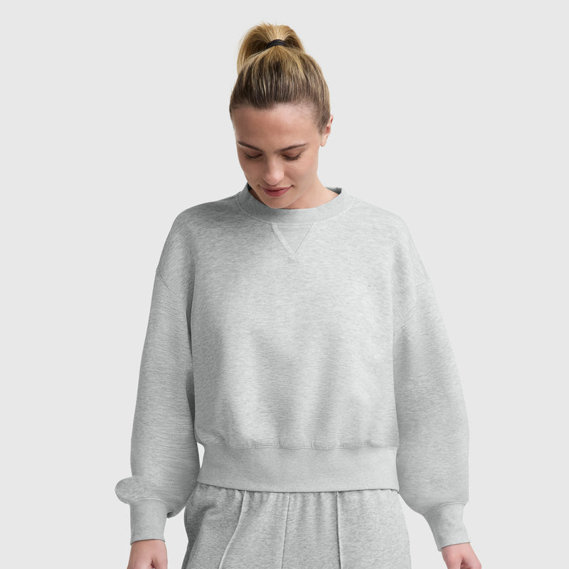 A woman with blonde hair in a ponytail wears a light gray Champion Endzone Loose Fit Crewneck, C Logo and matching sweatpants, looking down with a slight smile against a plain light background.