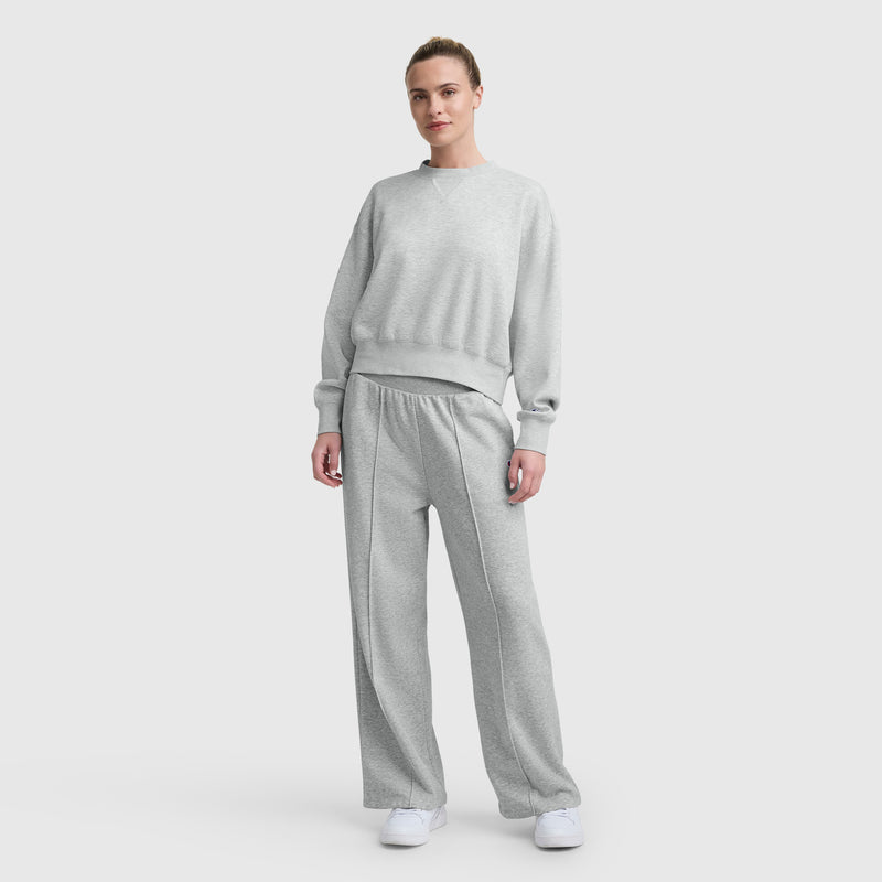 A person stands against a plain light background, wearing a light gray Champion Endzone Loose Fit Crewneck with C Logo in cozy scuba fabric, matching wide-leg sweatpants, and white sneakers for a comfortable, casual look.