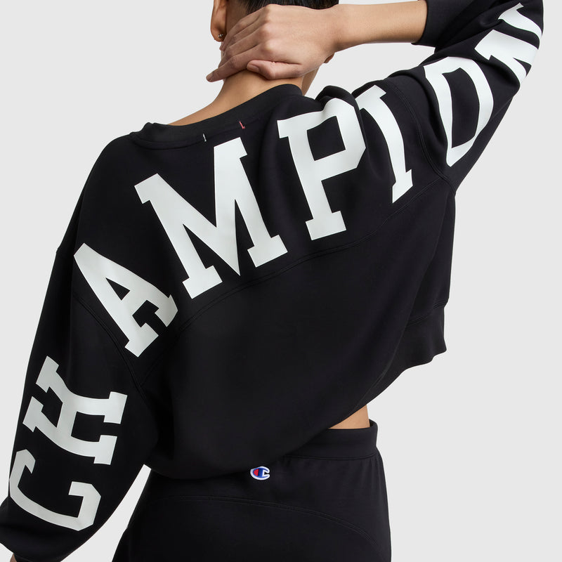 A person wears a black Champion Endzone Loose Fit Crewneck featuring the bold white C Logo and graphic detailing across the back and sleeve, with one hand resting on their head. Only the upper body is shown.