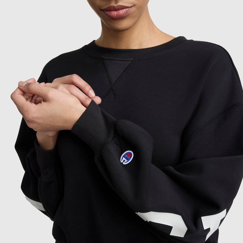 A person sports the Champion Endzone Loose Fit Crewneck, featuring a small C logo on the sleeve and white block numbers on the arm; only their lower face and upper torso are seen as they gently touch their own hand.