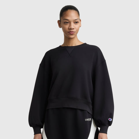 A woman with her hair pulled back wears the Champion Endzone Loose Fit Crewneck, featuring a C logo on the sleeve and lower left. The black sweatshirt’s balloon sleeves highlight its athletic heritage as she stands against a plain light background.