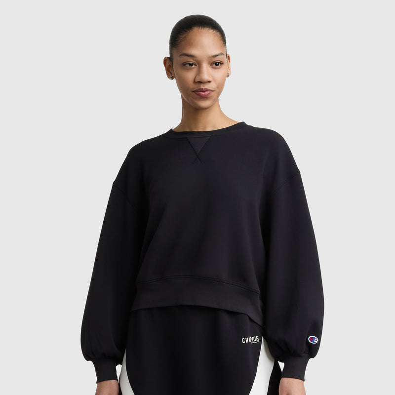 A woman with her hair pulled back wears the Champion Endzone Loose Fit Crewneck, featuring a C logo on the sleeve and lower left. The black sweatshirt’s balloon sleeves highlight its athletic heritage as she stands against a plain light background.