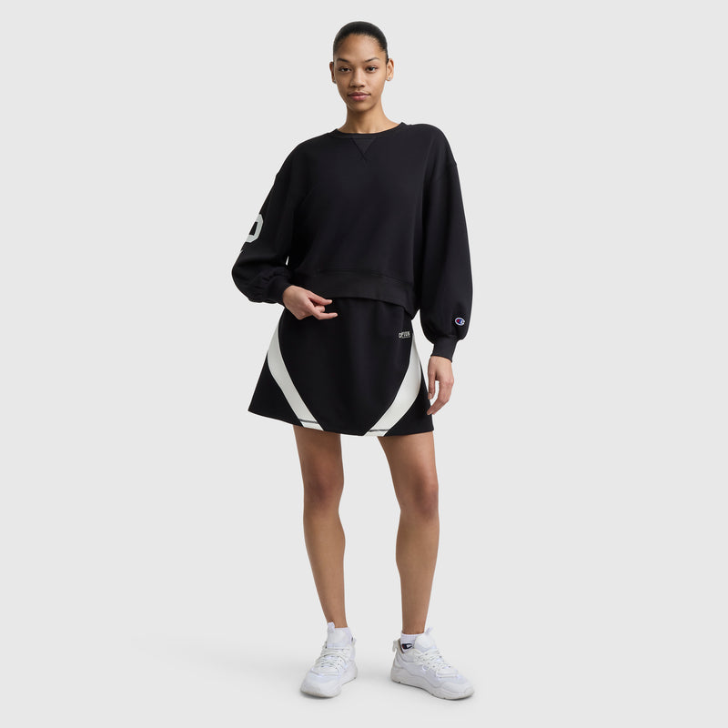A woman with her hair tied back poses against a plain background in a Champion Endzone Loose Fit Crewneck featuring a white C logo on the sleeve, paired with a black skirt with white stripes and white sneakers, reflecting Champion’s athletic heritage.