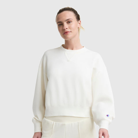 A woman with light hair tied back is wearing a Champion Endzone Loose Fit Crewneck, C Logo, styled as a women’s sport sweatshirt with matching white pants, standing against a plain light gray background.