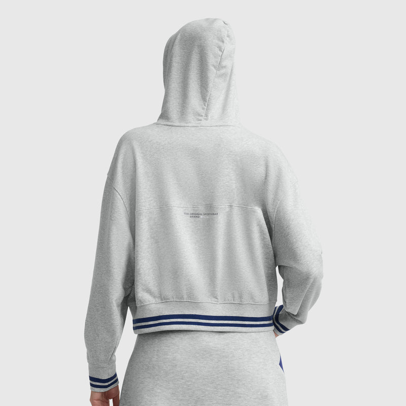 A person is seen from the back wearing the Champion Tackle Lace Front Hoodie in light gray with blue and white striped trim, hood up, featuring a tonal embroidered C logo at the center for a vintage sport-inspired look.