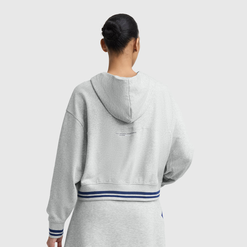 A person with dark hair in a bun is seen from behind wearing the Champion Tackle Lace Front Hoodie featuring a tonal embroidered C logo, with blue and white striped trim, against a plain light gray background.