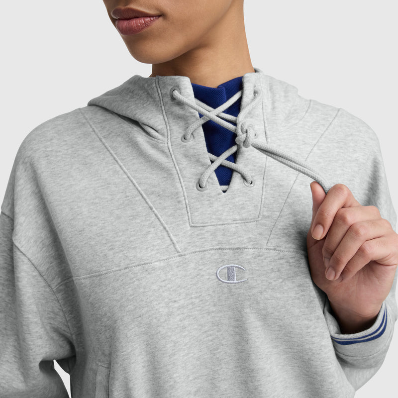 A person wears the Champion Tackle Lace Front Hoodie featuring a light gray color, lace-up neckline, and tonal embroidered C logo on the chest, highlighting classic sport-inspired style. One hoodie string is held in their hand.