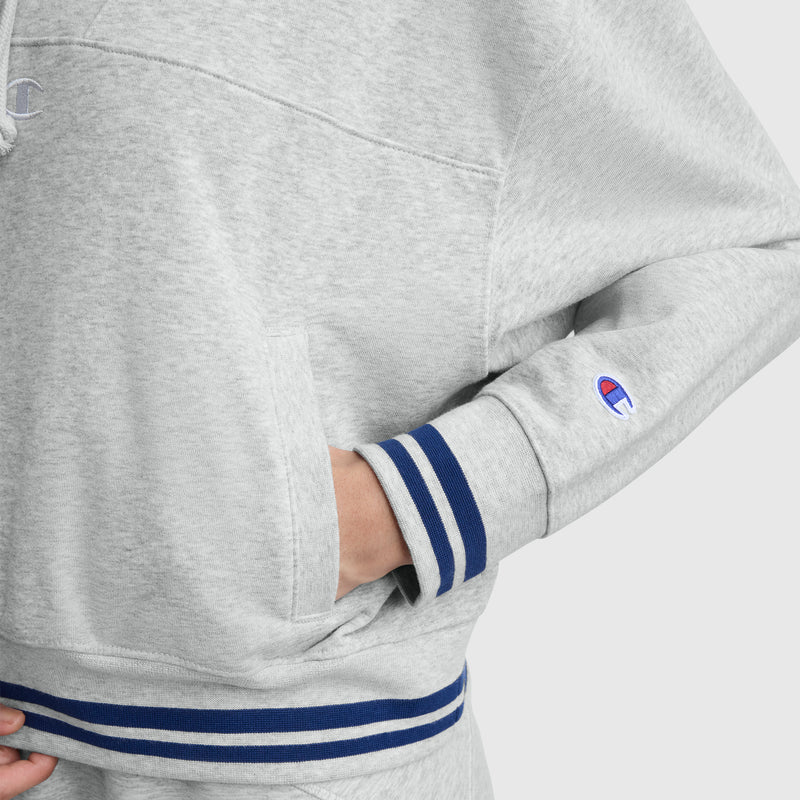 A person wears a light gray Champion Tackle Lace Front Hoodie with tonal embroidered C logo, featuring blue and white striped cuffs and hem, one hand tucked in the front pocket.
