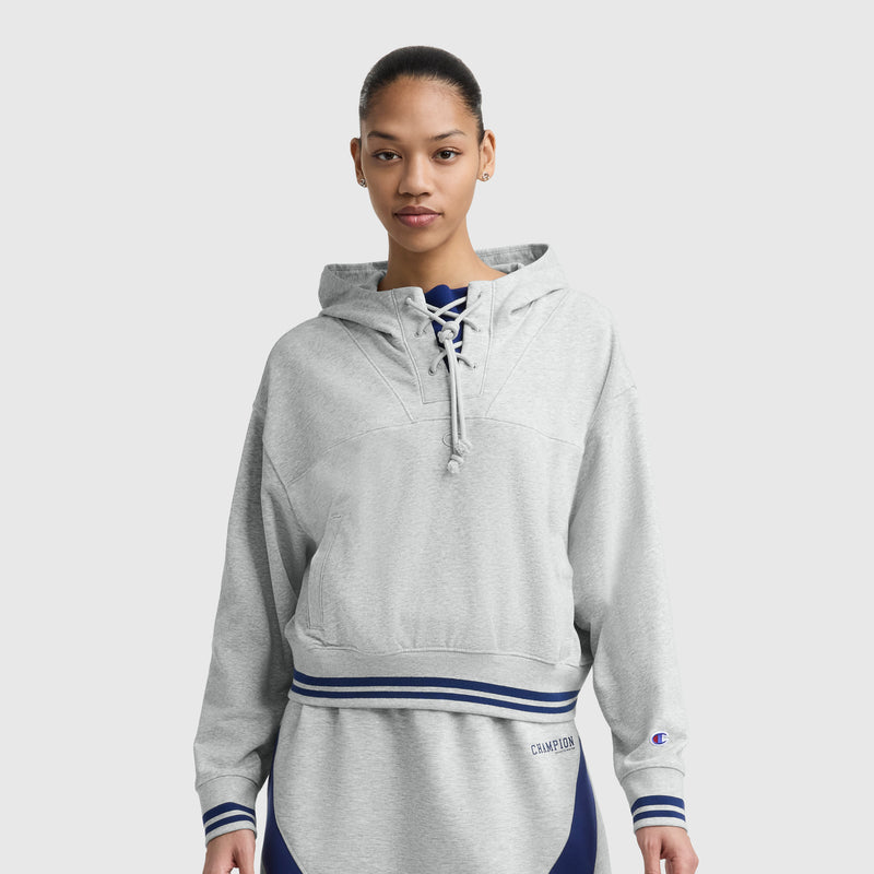 A woman faces forward in a light gray Champion Tackle Lace Front Hoodie with tonal embroidered C logo and lace-up collar, paired with matching sweatpants against a plain, light background.