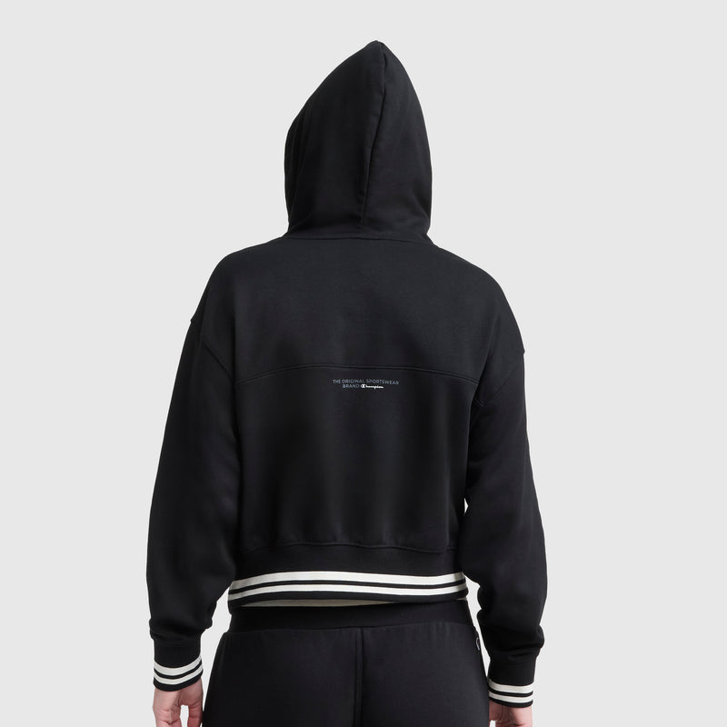 A person wearing a black Champion Tackle Lace Front Hoodie with a tonal embroidered C logo, featuring white striped cuffs and waistband. The hood is up, and there is small text on the upper back.