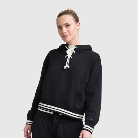 A person with light skin and blonde hair tied back is wearing the Champion Tackle Lace Front Hoodie, featuring a tonal embroidered C logo, white drawstrings, and striped cuffs and hem, standing against a plain light gray background.