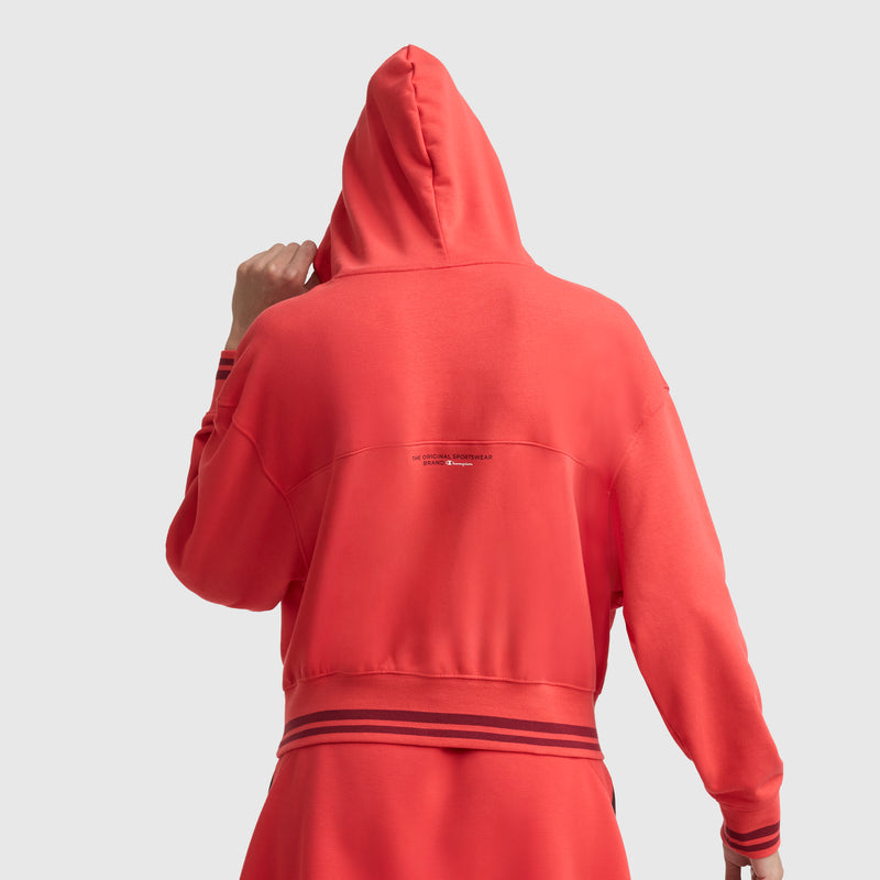 A person is seen from the back wearing Champion’s Tackle Lace Front Hoodie in bright red with the hood up. The tonal embroidered C logo and small text are on the upper back, finished with striped ribbed cuffs and a hem.