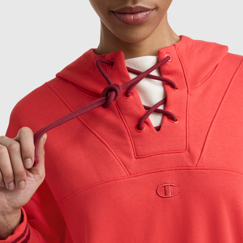 A person wears the Champion Tackle Lace Front Hoodie with a tonal embroidered C logo, holding a drawstring. The bright red hoodie partially reveals their mouth and chin, with the face only partly visible.