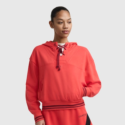 A young woman wearing a bright red hooded sweatshirt with a lace-up neckline and striped cuffs stands against a plain light gray background, looking to her left with one hand in her pocket.
