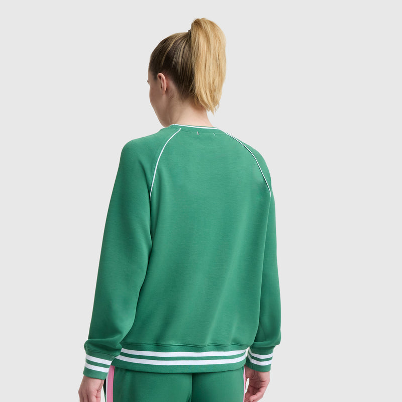 A blonde woman with a ponytail is seen from behind wearing the Champion Tennis Court Raglan Crewneck, C Logo, featuring white trim and matching green pants against a plain white background.