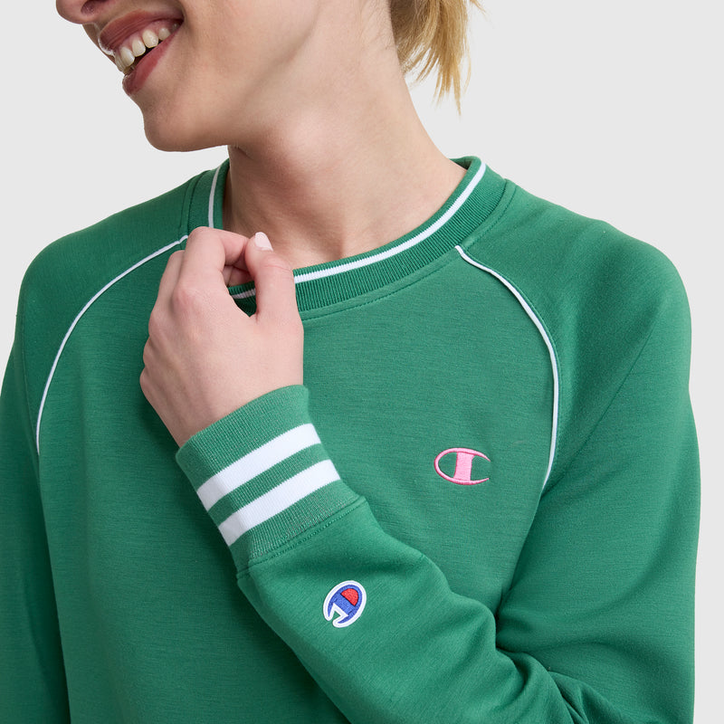 A woman smiles as she holds the collar of a green Champion Tennis Court Raglan Crewneck, C Logo, featuring white piping, striped cuffs, and Champion’s iconic logo on the chest and sleeve.