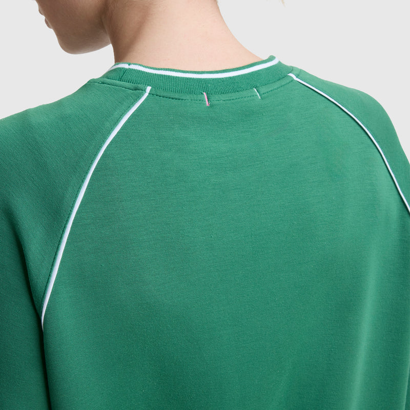 A close-up of a woman wearing the Champion Tennis Court Raglan Crewneck with white piping on the shoulders. The sporty C Logo sweatshirt is shown from the back, highlighting her neck and short hair.