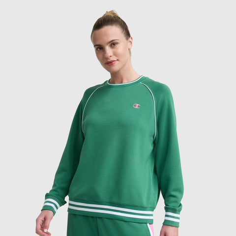 A blonde-haired woman in a bun wears the green Champion Tennis Court Raglan Crewneck, C Logo, and matching sweatpants. The sweatshirt has white stripes on the collar, cuffs, and hem with a small embroidered C logo. The background is plain white.