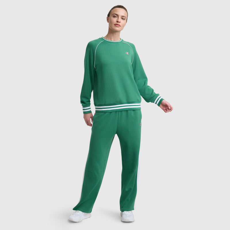 A person stands against a plain background wearing the Champion Tennis Court Raglan Crewneck, C Logo sweatshirt in green with matching sweatpants featuring white stripes, paired with white sneakers.