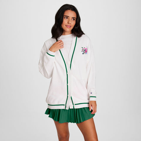 A woman wears a Champion Classic T-Shirt with a script logo under a white cardigan and green pleated skirt, standing against a plain white background and looking slightly to the side.