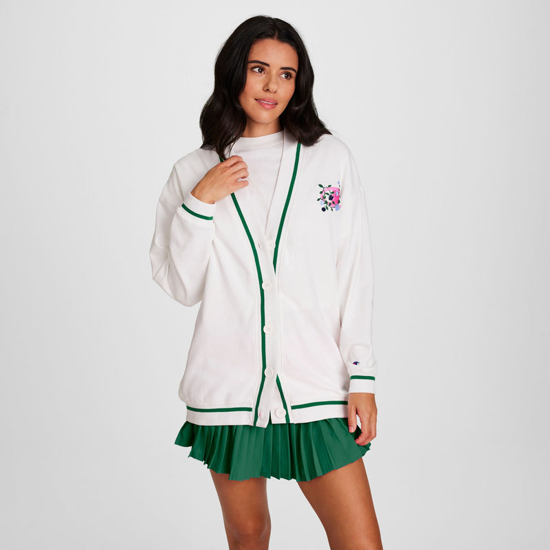 A woman wears a Champion Classic T-Shirt with a script logo under a white cardigan and green pleated skirt, standing against a plain white background and looking slightly to the side.