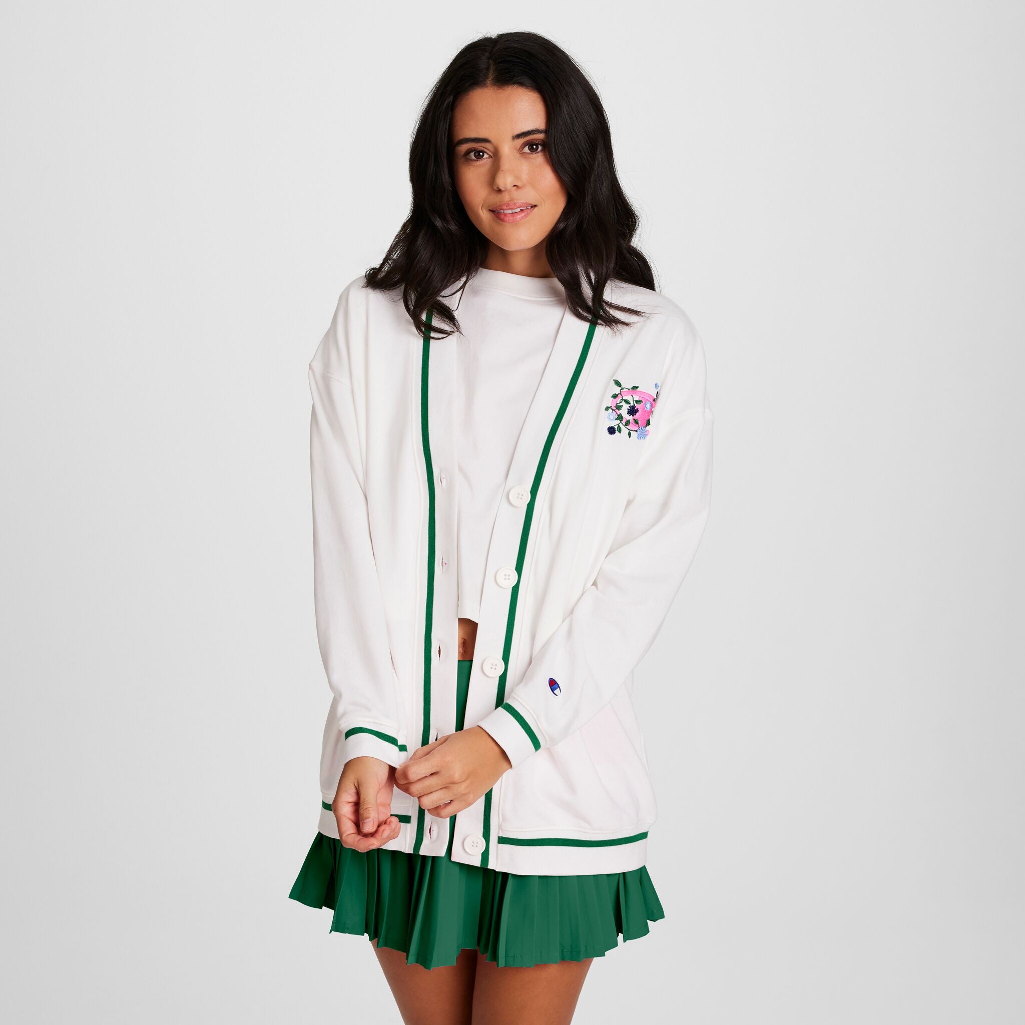 A woman with long dark hair wears a white cardigan with green trim over the Champion Classic T-Shirt, Script Logo, paired with a green pleated skirt. She stands against a plain white background, looking at the camera and holding her cardigan.
