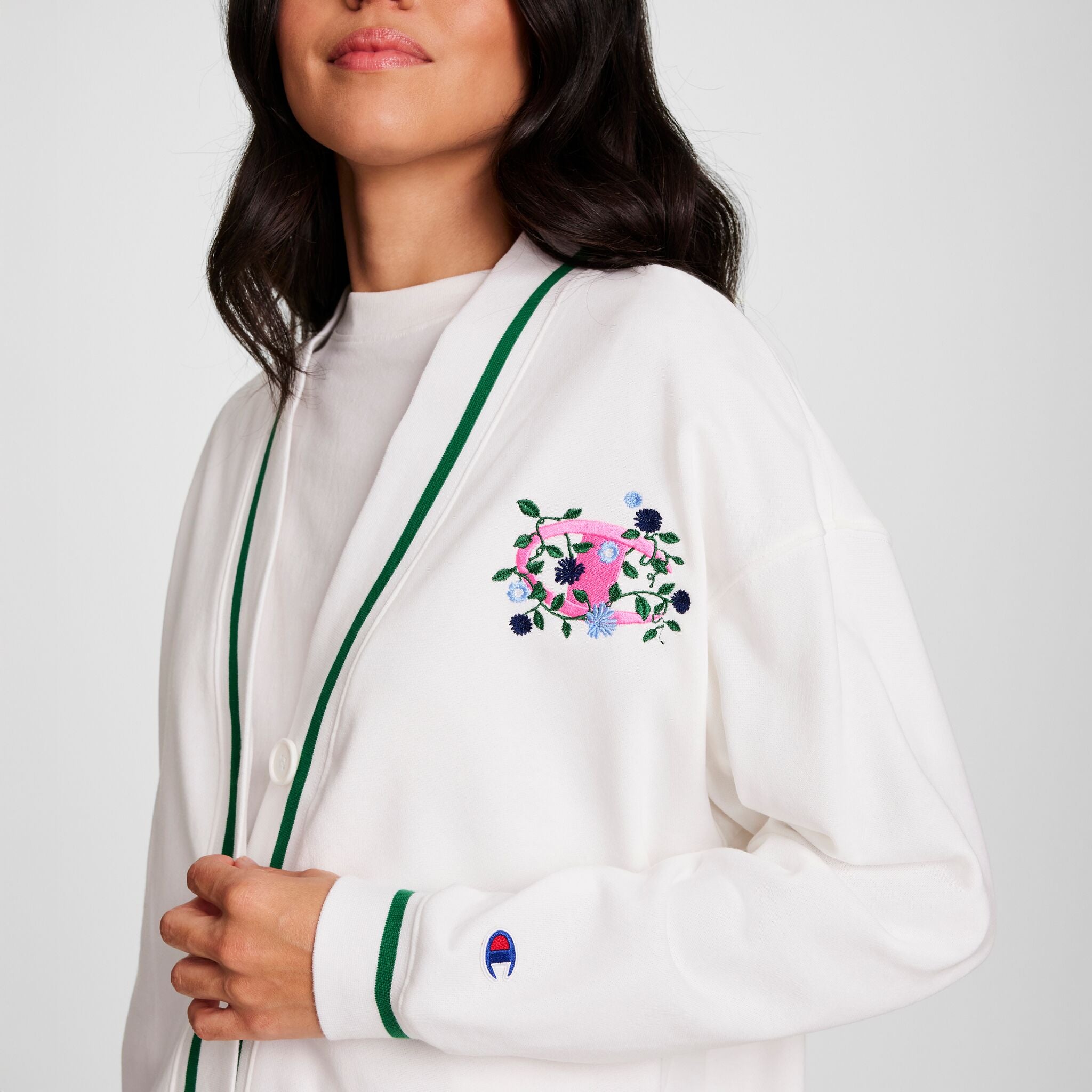 A woman with long dark hair stands against a plain background wearing a white cardigan with green trim and an embroidered pink cup layered over the Champion Classic T-Shirt with Script Logo.