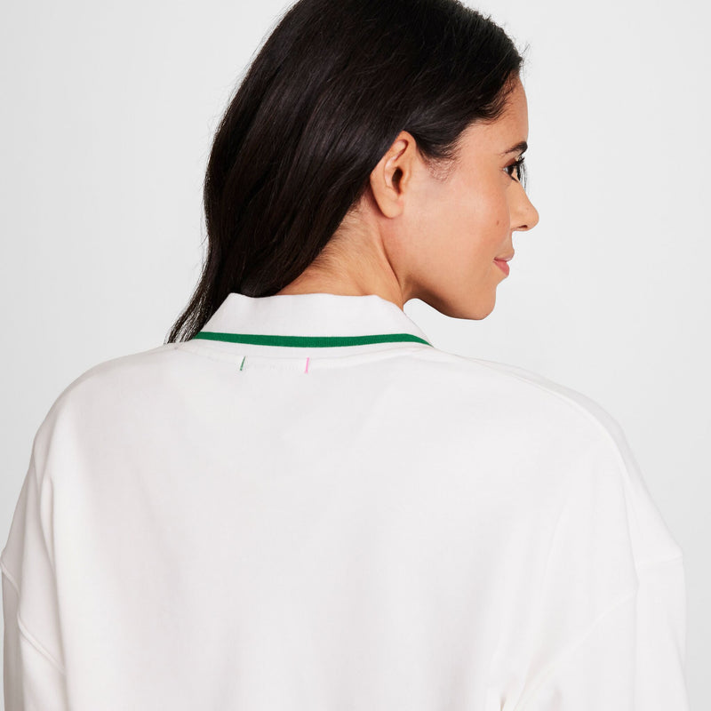 A woman with long dark hair, seen from behind and looking over her shoulder, wears a Champion Classic T-Shirt, Script Logo—white with a green collar stripe—against a plain light background.