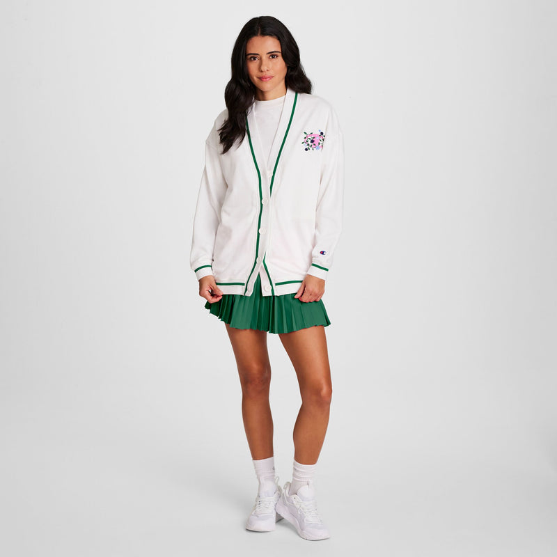 A woman with long, dark hair poses casually against a plain background, wearing a Champion Classic T-Shirt with Script Logo beneath a white cardigan trimmed in green, paired with a green pleated skirt, white socks, and sneakers.