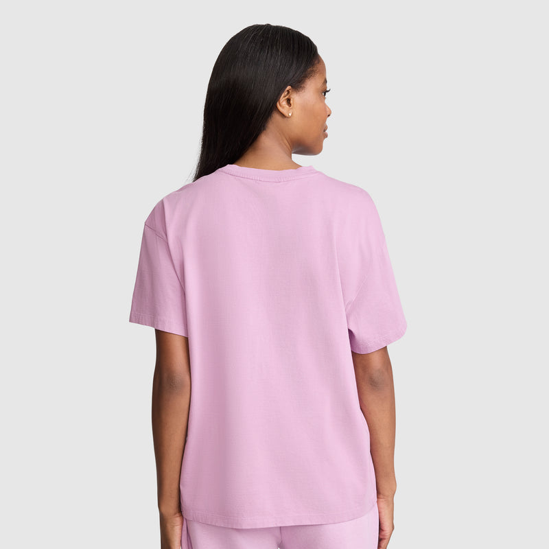 A person with long dark hair is standing facing away, wearing the Champion Short Sleeve Loose Fit T-Shirt in light pink and matching pants against a plain light gray background.