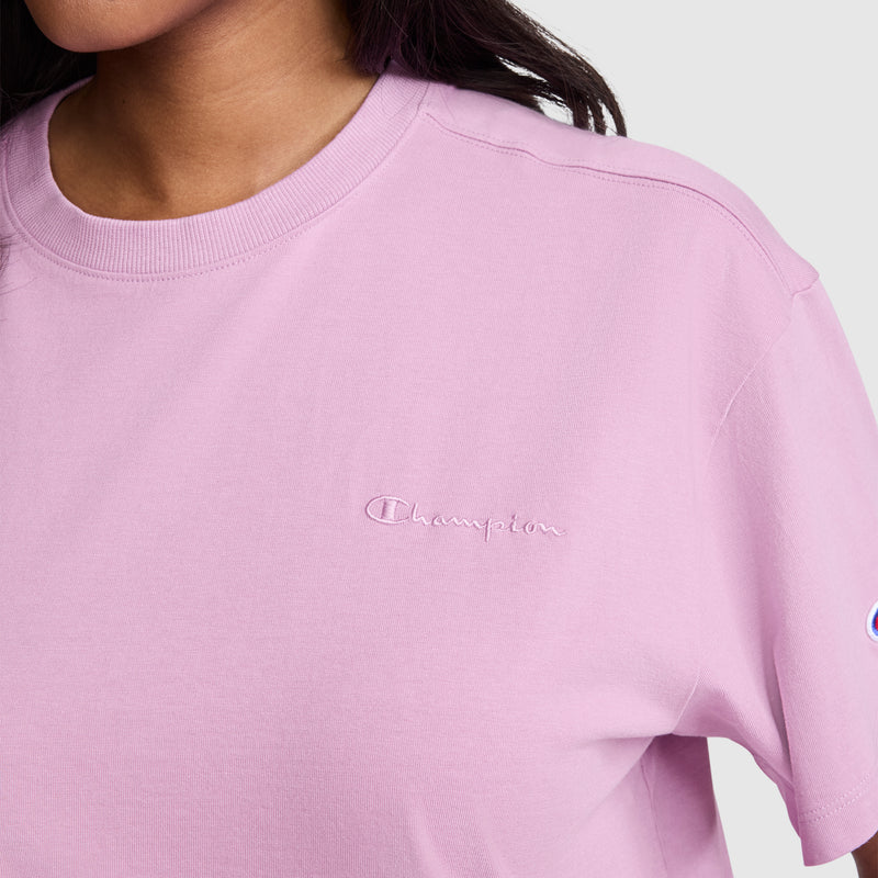 A person wearing the Champion Short Sleeve Loose Fit T-Shirt in light purple, featuring an embroidered Champion logo on the chest and a partially visible logo patch on the sleeve. Made from comfortable cotton jersey material, this women's t-shirt provides a relaxed fit for casual style and all-day comfort.