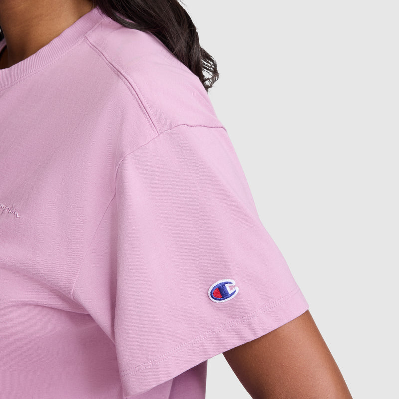 A person is shown from the upper body and part of the arm, wearing the Champion Short Sleeve Loose Fit T-Shirt in light pink. The soft cotton jersey tee features the Champion logo on the sleeve and is set against a plain background.