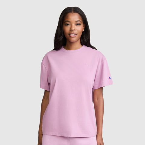 A woman with long dark hair wears the Champion Short Sleeve Loose Fit T-Shirt and matching light purple shorts, standing and smiling against a plain light gray background.