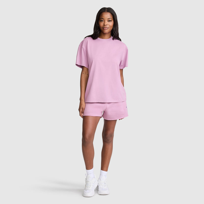 A woman stands against a plain background wearing the Champion Short Sleeve Loose Fit T-Shirt in pink, paired with matching shorts, white socks, and white sneakers. Made from soft cotton jersey for all-day comfort, the t-shirt offers a relaxed fit. She is smiling with her arms relaxed by her sides.