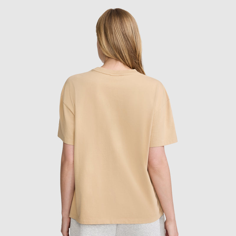 A person with straight, light brown hair is seen from the back against a plain, light background, wearing a beige Champion Short Sleeve Loose Fit T-Shirt and light gray pants.