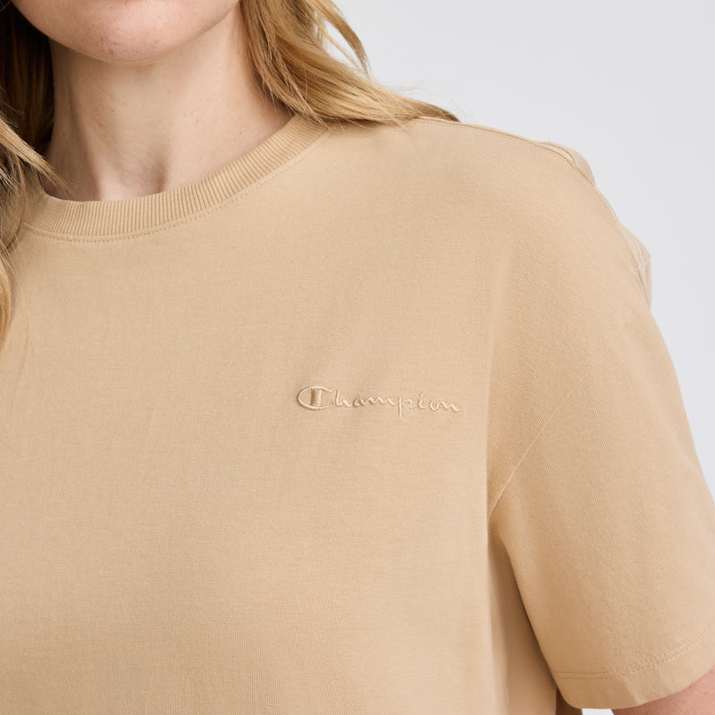 A person wearing a beige Champion Short Sleeve Loose Fit T-Shirt, made from soft cotton jersey with an embroidered logo on the chest. Only the lower face, neck, and upper torso are visible; hair is blonde and wavy.