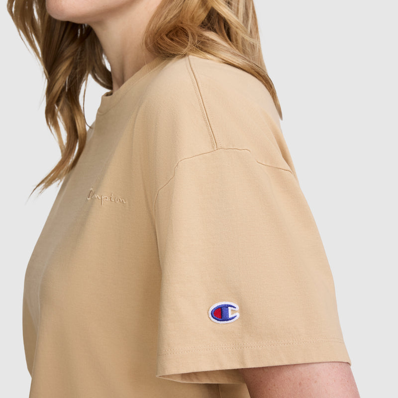 A person shown from the side against a plain background wears the Champion Short Sleeve Loose Fit T-Shirt in beige, made from soft cotton jersey, featuring the Champion logo embroidered on the sleeve and "Champion" stitched on the chest.