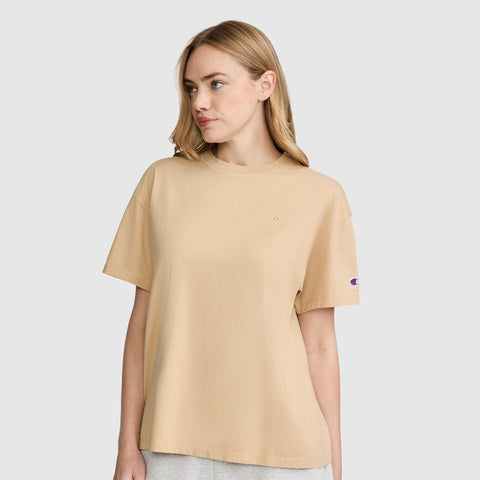 A woman with light brown hair wears the Champion Short Sleeve Loose Fit T-Shirt in beige and light gray pants, standing against a plain light background while looking to her left.