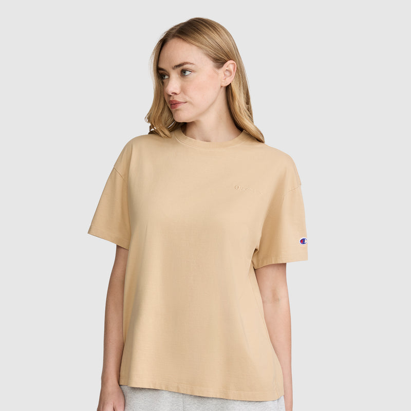 A woman with light brown hair wears the Champion Short Sleeve Loose Fit T-Shirt in beige and light gray pants, standing against a plain light background while looking to her left.